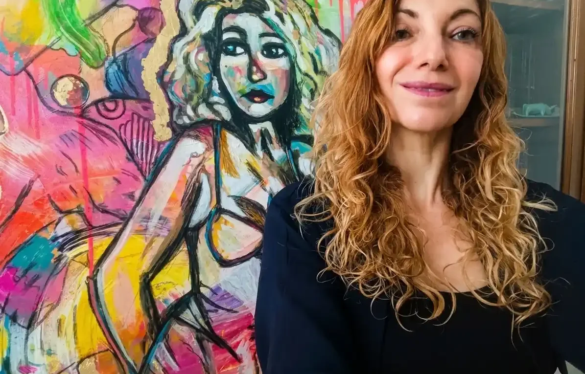 Une femme souriante aux cheveux bouclés posant devant une peinture expressionniste colorée, illustrant son rôle de coach des artistes.
