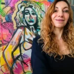 Une femme souriante aux cheveux bouclés posant devant une peinture expressionniste colorée, illustrant son rôle de coach des artistes.