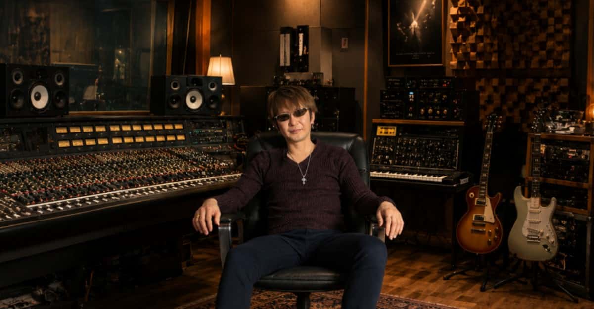 Un portrait de nobutaka soeda assis dans un studio d'enregistrement professionnel, devant une grande console de mixage analogique entourée d'équipements audio vintage et de guitares électriques.