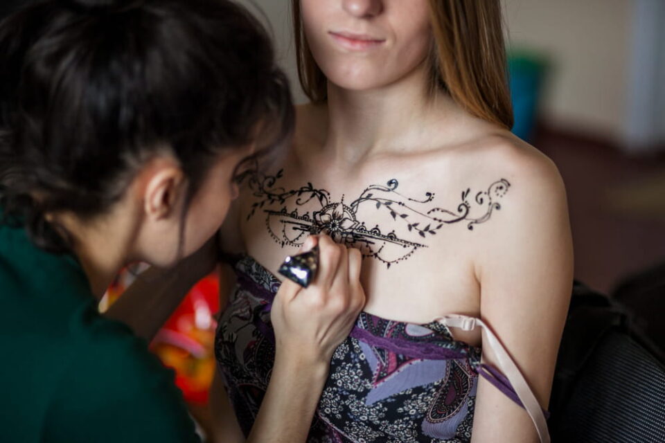 Séance de tatouage au henné sur le décolleté, avec motifs floraux complexes en cours de création.