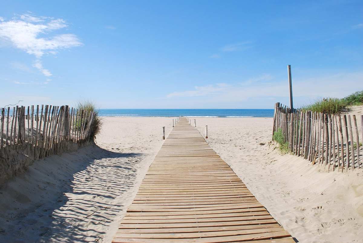 Passerelle en bois sur la plage, bordée de clôtures, menant à la mer sous un ciel bleu Passerelle en bois sur la plage, bordée de clôtures, menant à la mer sous un ciel bleu